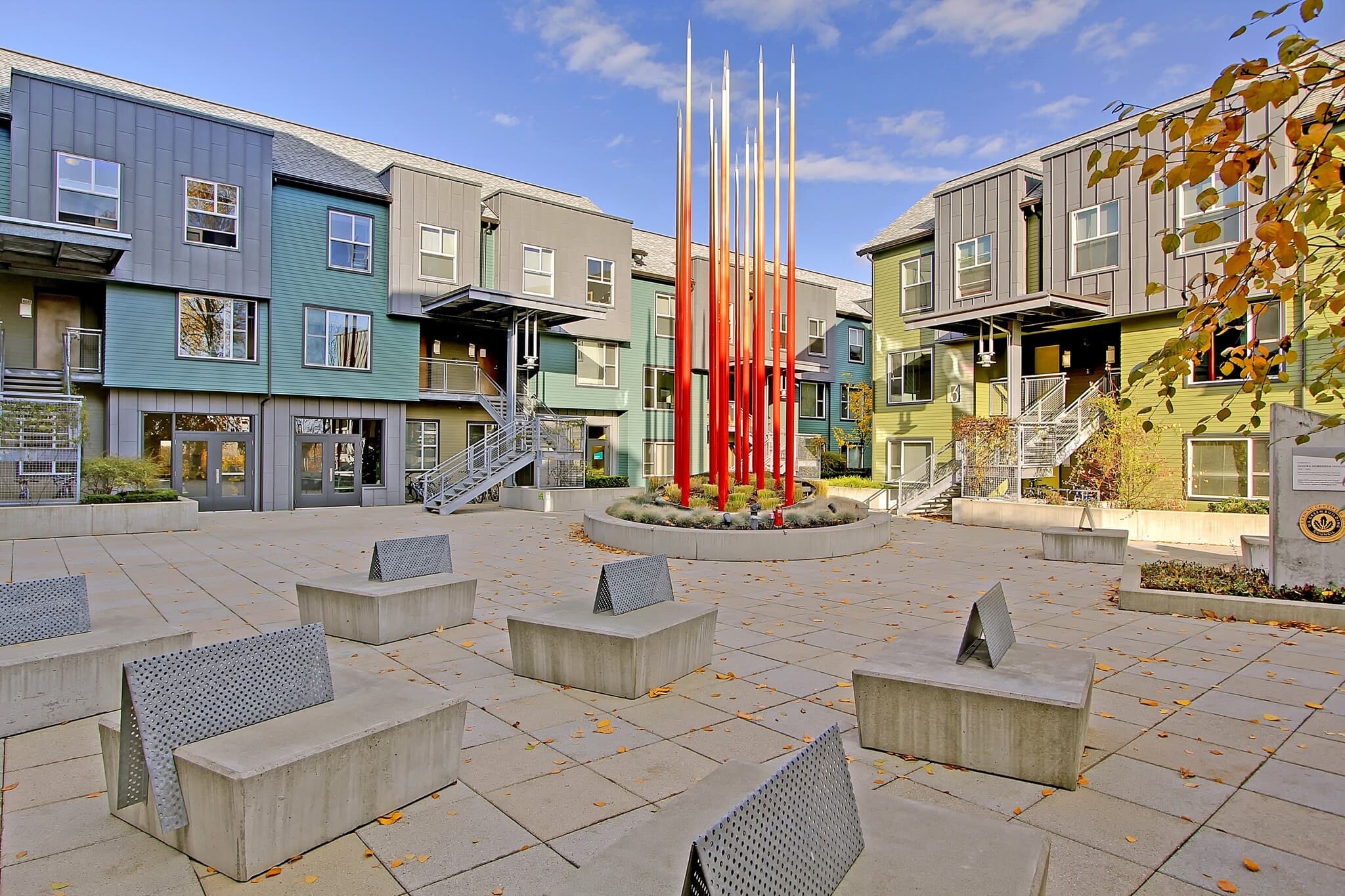 Modern Nordheim Court student housing features a courtyard with geometric concrete benches, a circular planter, and tall red metal poles as art. Green and gray UW student apartments with balconies enclose the open space under a blue sky.