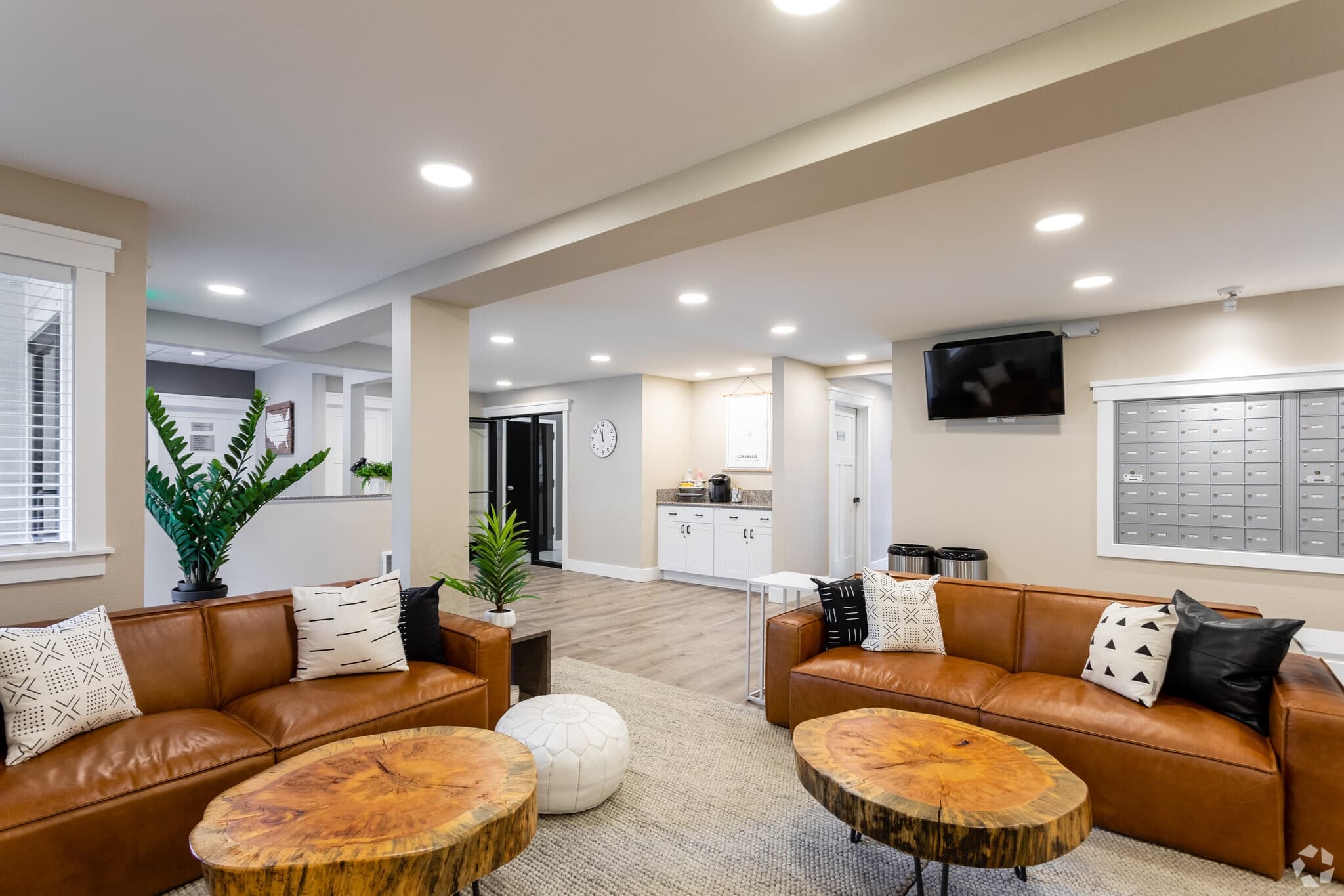 Modern apartment lounge at Oak Vale Apartments with two brown leather sofas, wooden coffee tables, decorative plants, wall-mounted TV, mailboxes, and a kitchenette. Neutral tones and recessed lighting create a cozy atmosphere.