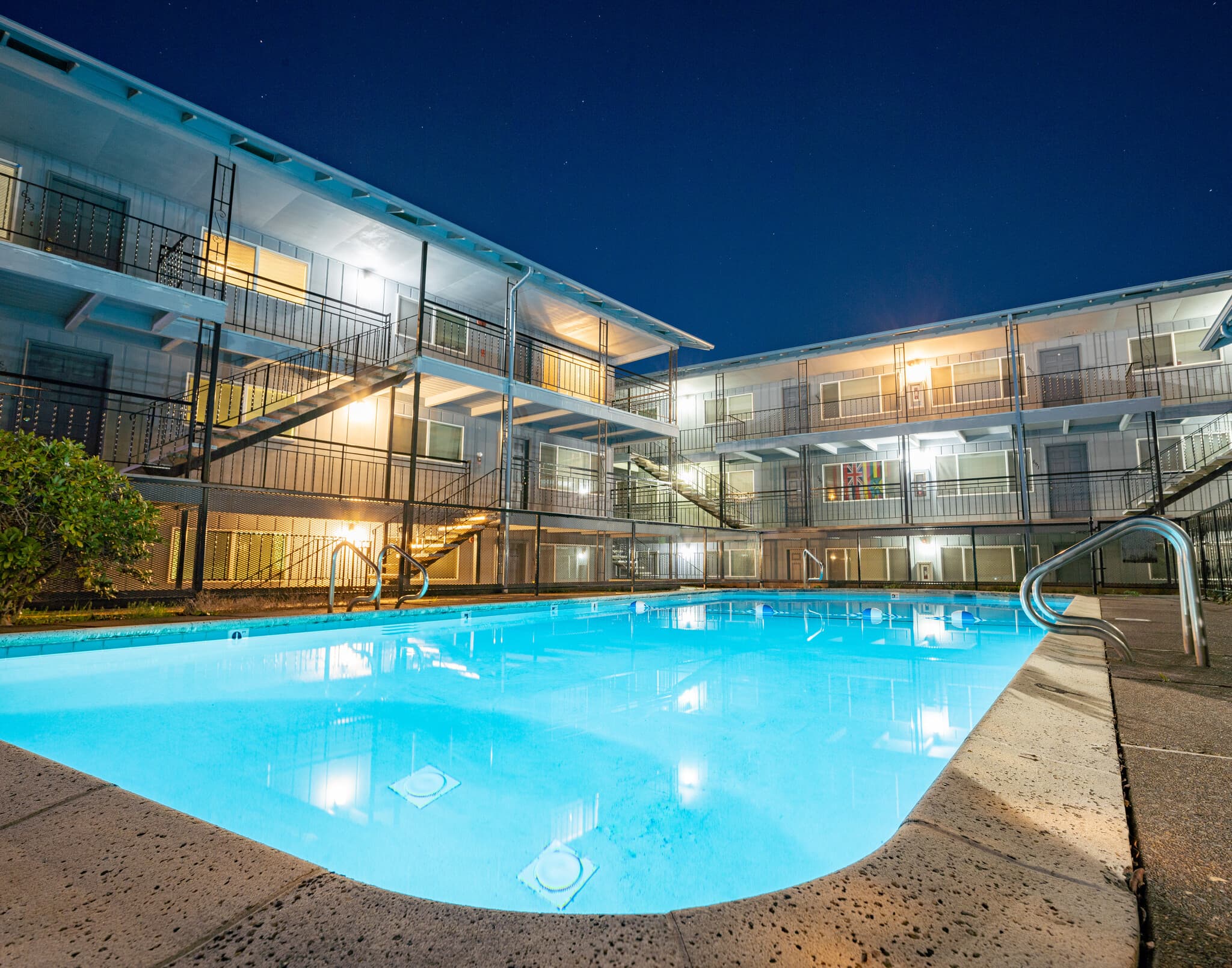 A brightly lit outdoor swimming pool in the vicinity of a three-story motel building at night, with illuminated balconies and stairways reflecting on the water.