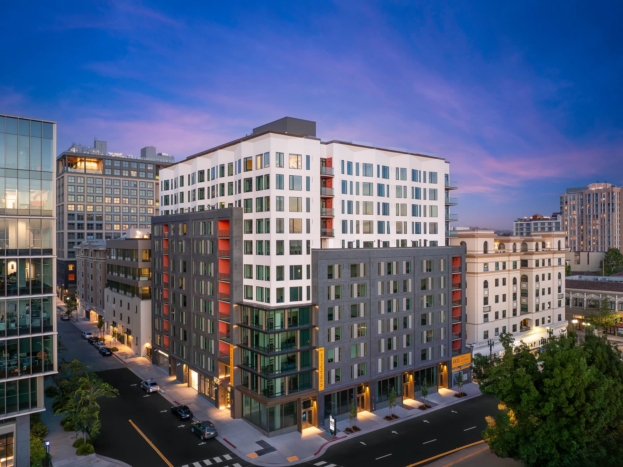 Ace Berkeley, a modern mid-rise apartment building with a white and dark gray exterior, stands at a city intersection during sunset, its illuminated windows shining against the urban backdrop.