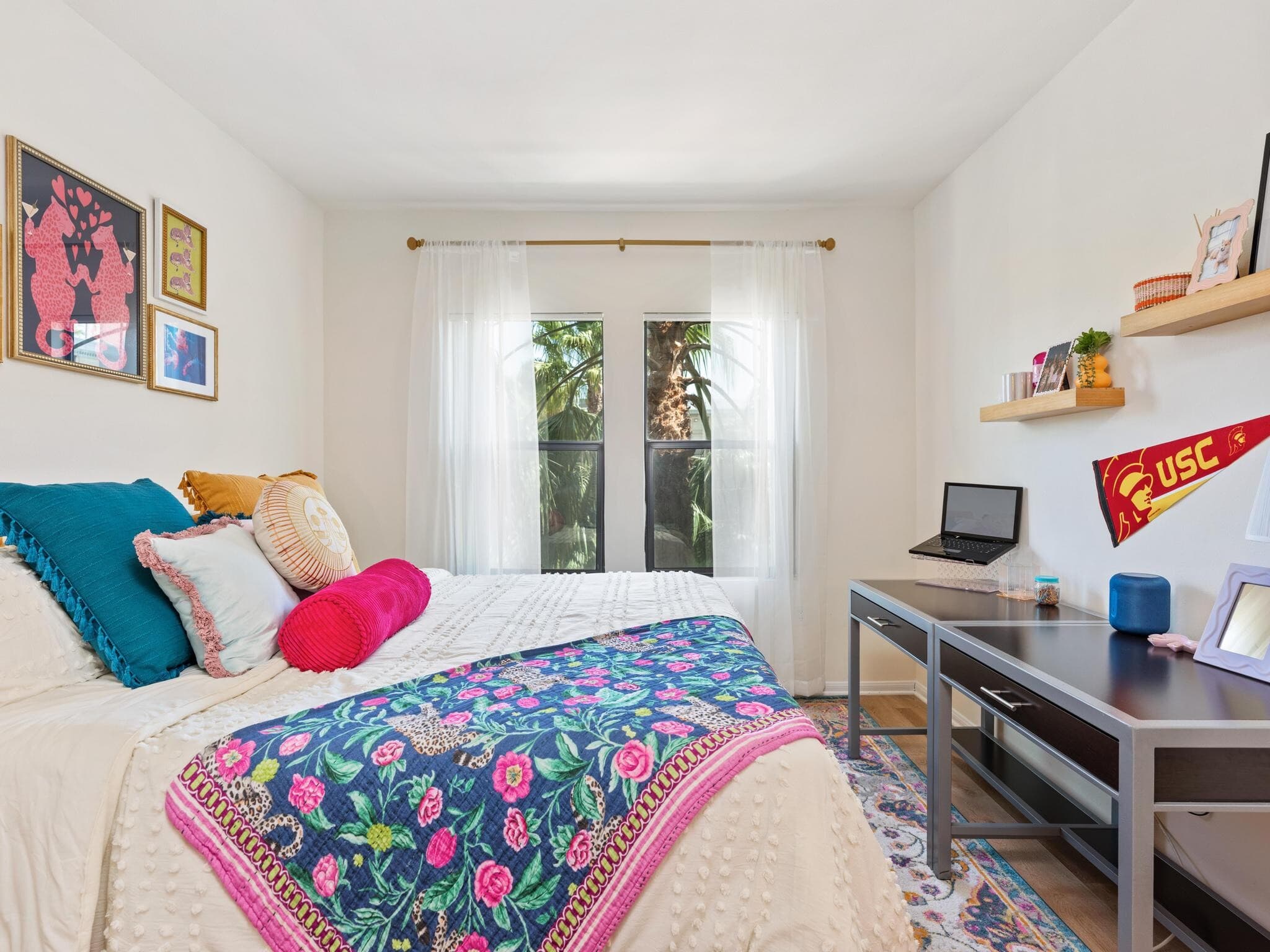 A bright bedroom with a bed covered in colorful pillows and a floral blanket, artwork on the walls, a desk with a laptop, and sheer curtains on windows letting in natural light with palm trees visible outside.