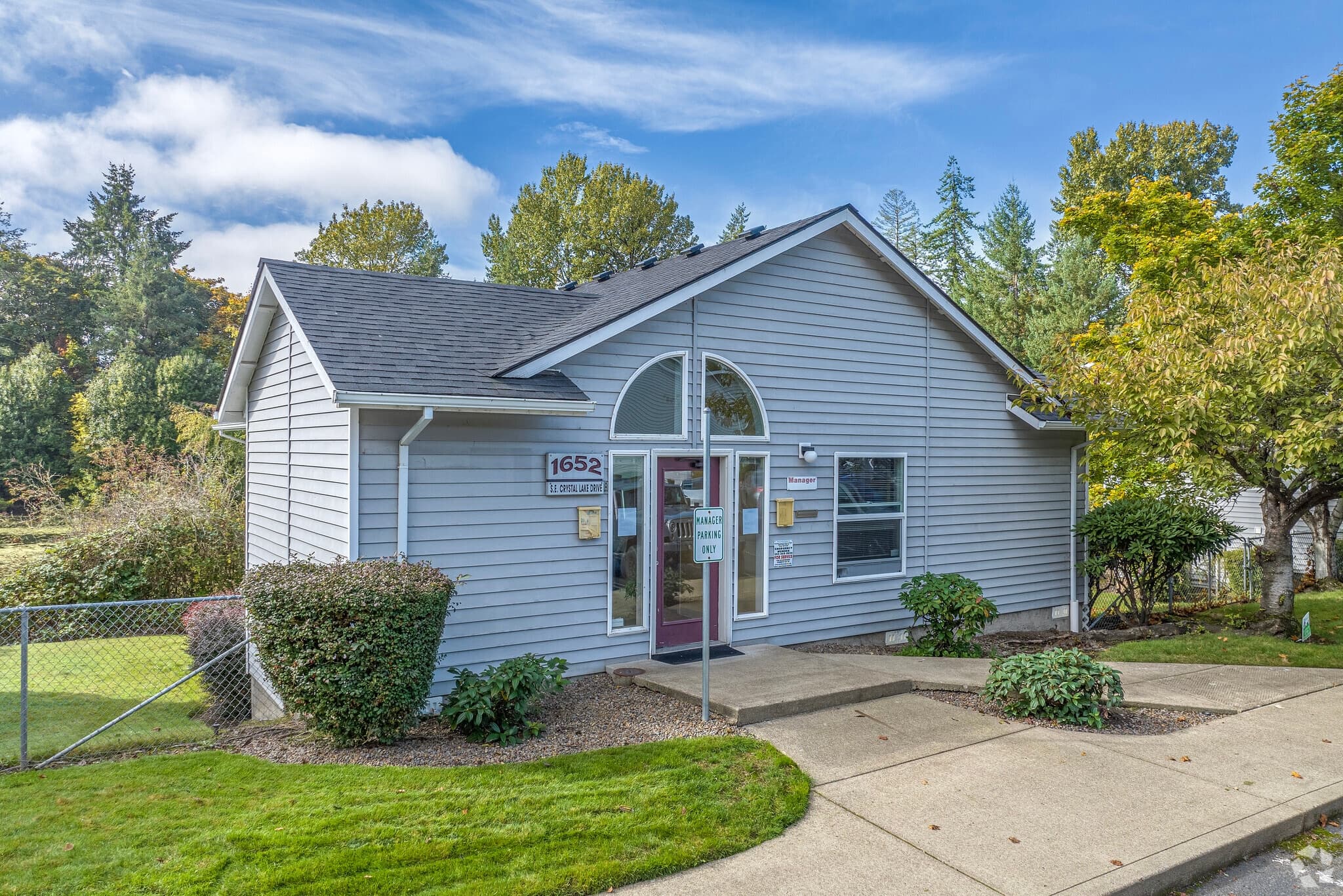 A small, light gray building with a gabled roof, glass front door, and address number 1652 sits at Crystal Lake Apartments. It's surrounded by green grass, shrubs, trees, a sidewalk, and a chain-link fence under a partly cloudy sky.