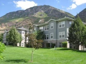 Gray Harris Condo buildings surrounded by green lawns and trees, with mountains and a partly cloudy blue sky in the background.