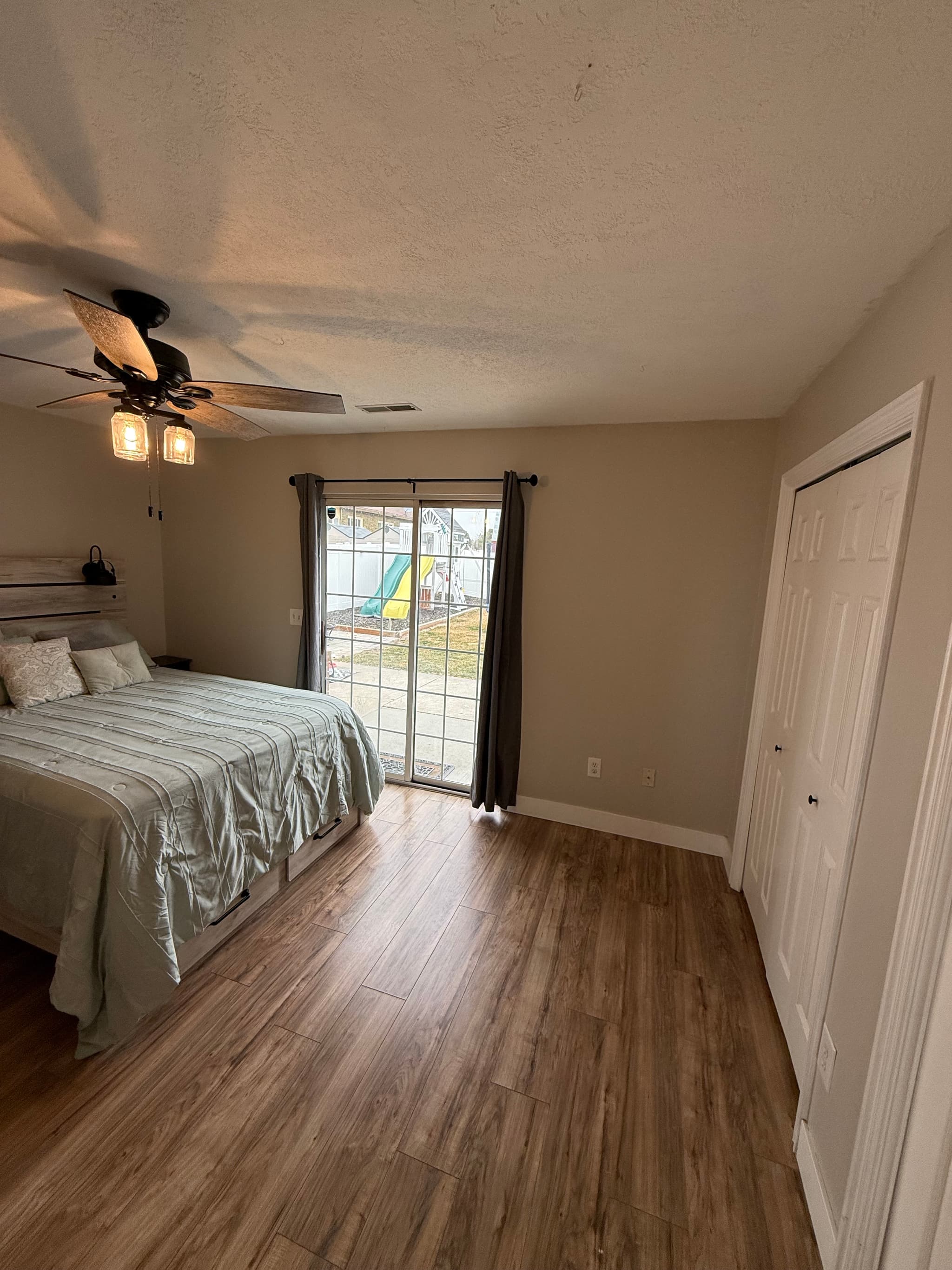 A cozy apartment bedroom at 282 W 960 S features wood flooring, a bed with pillows and blankets, ceiling fan, sliding glass doors with gray curtains overlooking a playground, and a white double-door closet.