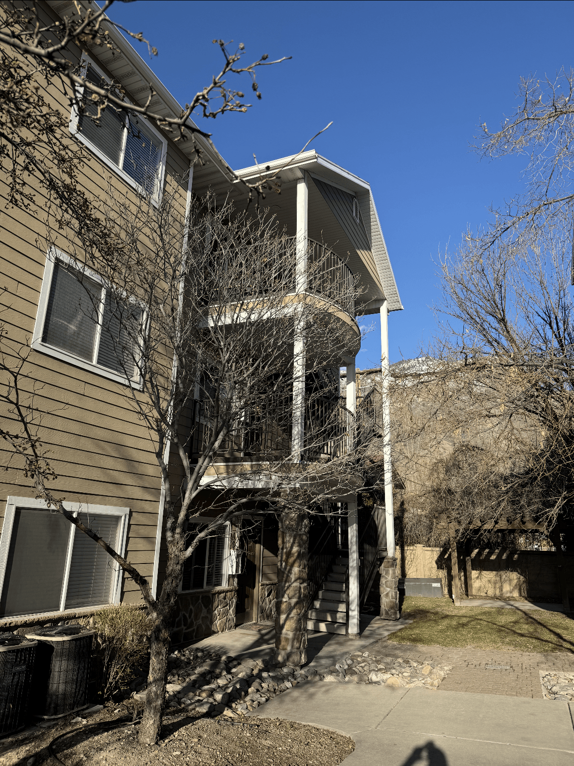 A three-story apartment building at 389 S State St, Apt 25, with beige siding and white trim is shown on a sunny day. Leafless trees stand in front, and a staircase leads to an upper porch. Shadows fall across the sidewalk and building.