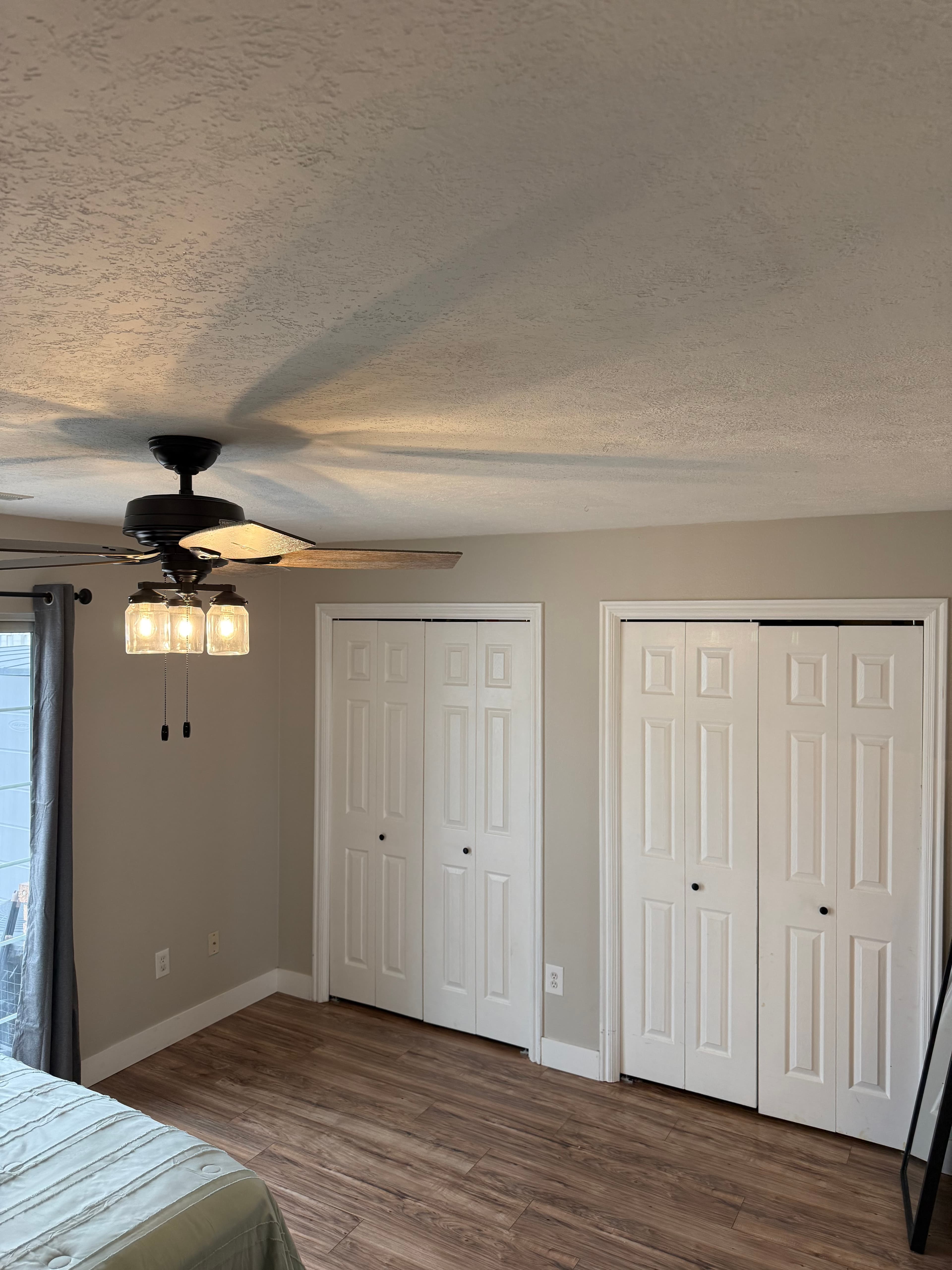 A bedroom in this apartment at 282 W 960 S features light brown wood flooring, two sets of white double-door closets, gray walls, a ceiling fan with three lights, and a window with gray curtains on the left.