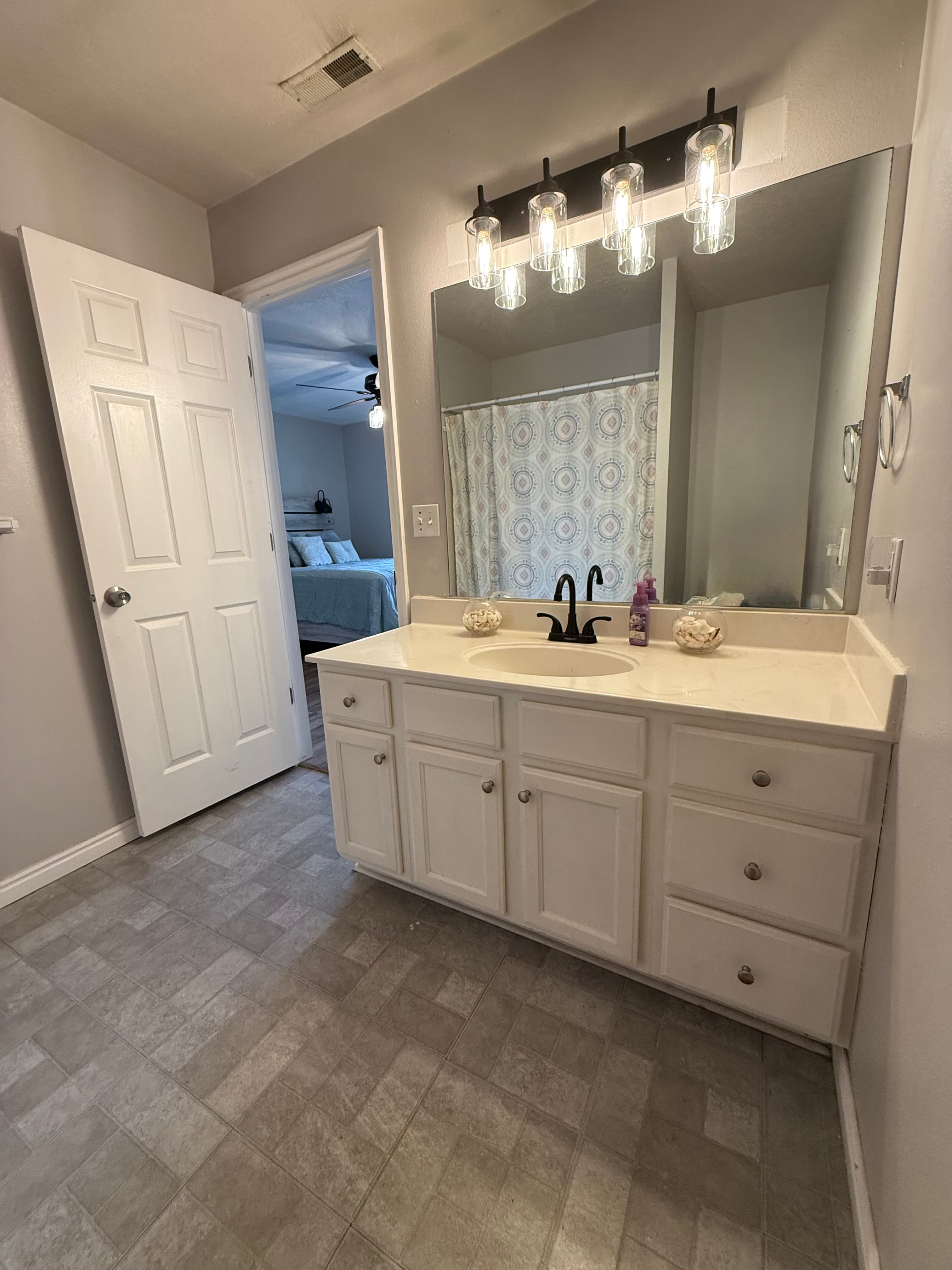 A bathroom at 282 W 960 S, APT C features a white vanity, black faucet, large mirror, and four-bulb light fixture. Tiled floors lead to a shower with a patterned curtain and an open door revealing the bedroom in the background.