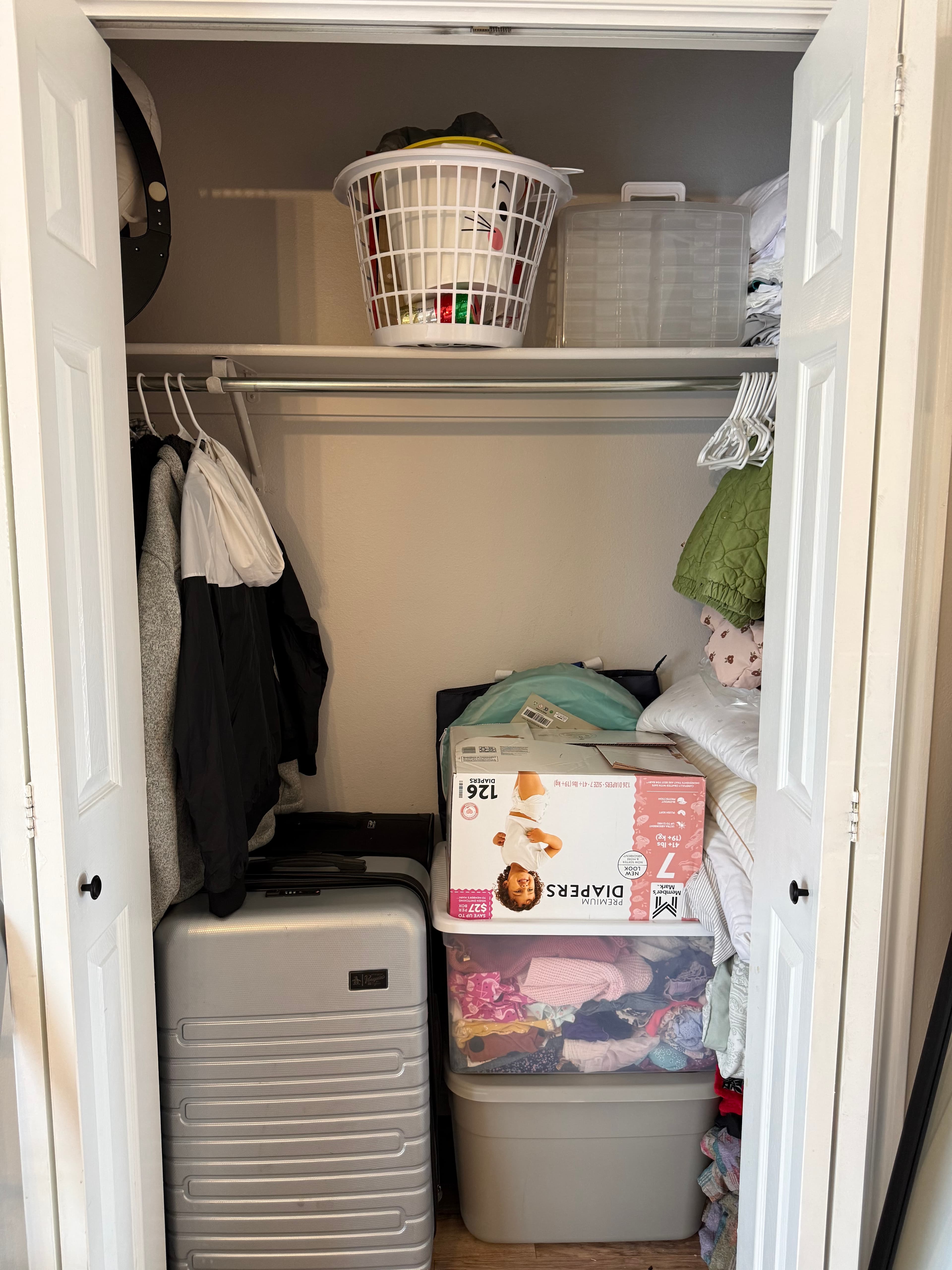 A small apartment closet at 282 W 960 S features white folding doors, hanging clothes, a suitcase, a plastic bin with folded clothes, a box of diapers, a laundry basket, and various items neatly stored on shelves and the floor.