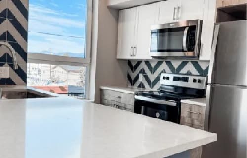 Modern kitchen at Jefferson Flats with white cabinets, stainless steel appliances, microwave, oven, and refrigerator. A white countertop island and geometric blue-and-white tile backsplash brighten these stylish apartments. Large window lets in natural light.