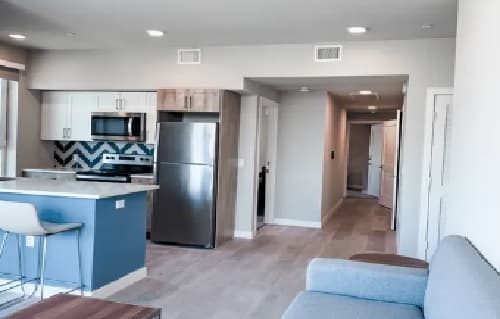 Modern apartment interior at Jefferson Flats with an open kitchen featuring white cabinets, stainless steel appliances, blue accent backsplash, and a breakfast bar. A hallway extends to the back, with part of a gray sofa visible in the foreground.