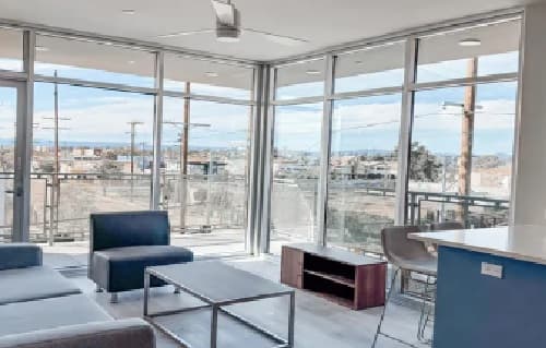 Modern living room at Jefferson Flats with floor-to-ceiling windows, gray sofas, a coffee table, and a small TV stand. Step onto the balcony to enjoy cityscape views under a bright blue sky with scattered clouds.