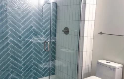 Modern bathroom at Jefferson Flats with a glass-enclosed shower featuring blue herringbone tile on one wall, white rectangular tiles on another, a metal showerhead, and a white toilet next to a towel bar.