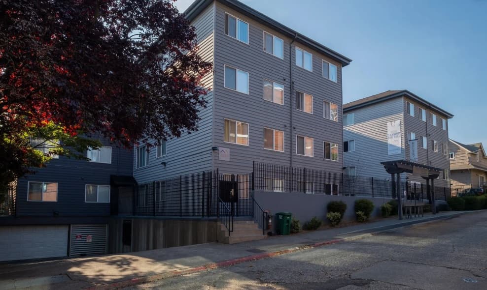 Kelsey Apartments is a modern, four-story gray building offering U-District student housing with many windows, a gated entry, and garage doors at street level, situated on a sloped road with trees and shrubs nearby.