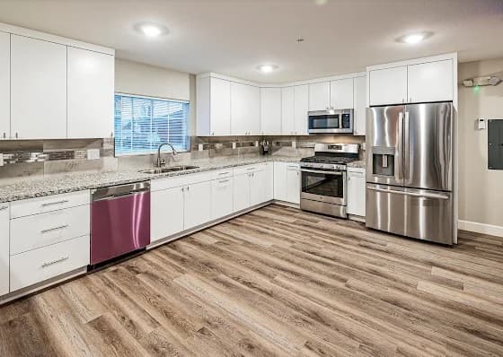 Modern kitchen in Kramer's House Downtown Reno features white cabinets, stainless steel appliances, granite countertops, a wood-style floor, and a large window with blinds above the sink. The room is well-lit with recessed lighting.