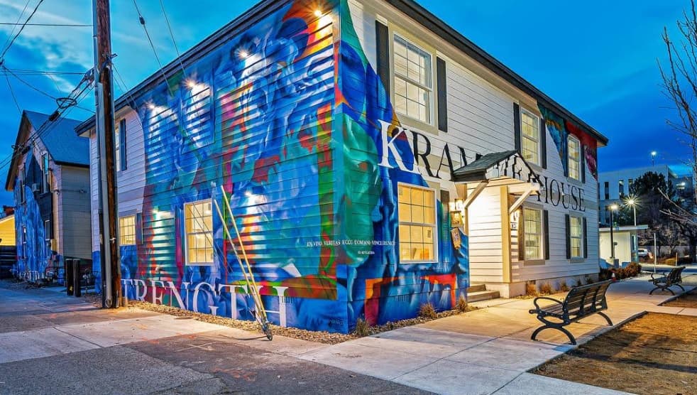 A two-story white building in Kramer's House Downtown Reno, covered in a colorful mural with abstract faces and the word STRENGTH, illuminated by lights at dusk, with benches and a sidewalk in front.