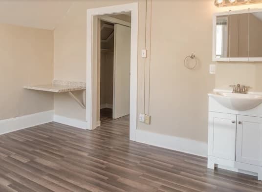 A room in Kramer's House Downtown Reno features wood-style flooring, beige walls, a small white sink with mirror, a wall-mounted countertop, and an open doorway leading to a walk-in closet.