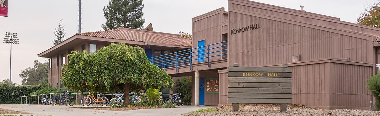 A two-story building labeled “Konkoly Hall” with a row of bicycles parked outside, a wooden sign in front, and trees and shrubs surrounding the area.