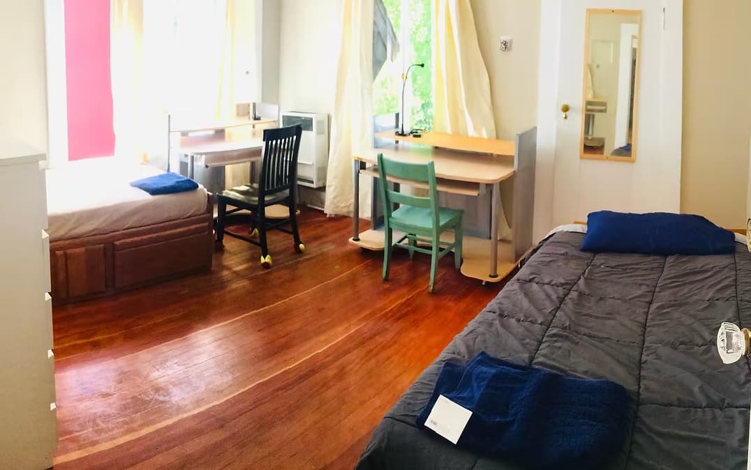 A bright, tidy student housing bedroom in Kluegel House features two single beds with dark bedding and towels, two desks by the window, hardwood floors, and a wall mirror on the door. Sunlight streams in through the windows.