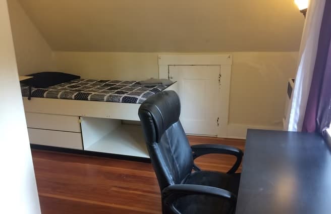 A cozy attic bedroom in Kluegel House, perfect for student accommodation, with wooden floors, a single bed with storage drawers, a desk and black office chair by the window, plus a small white door set into the sloped wall.