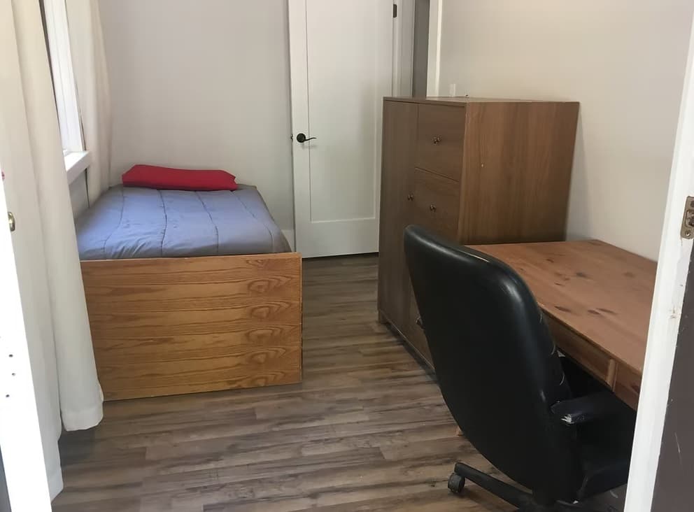 A small, simple bedroom in Kluegel House features a single bed with gray bedding and a red pillow, a wooden dresser, and a wooden desk with a black office chair—all ideal for student housing on a light wood floor.