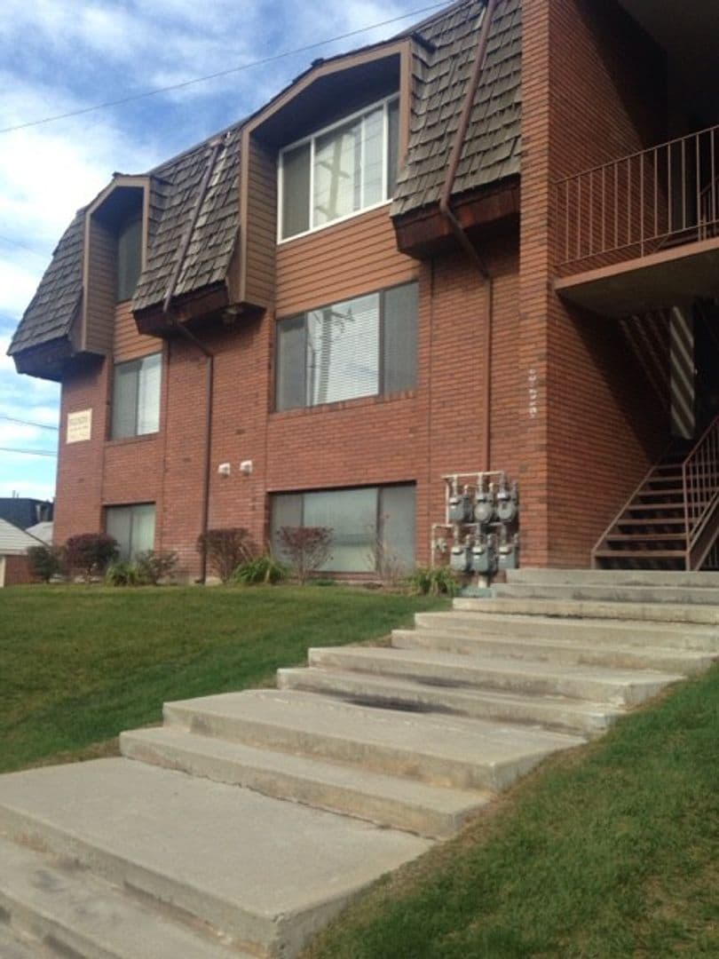 A two-story brick apartment building, known as Wellington Condos by Joseph Thomas, features brown shingle siding, large windows, an outdoor staircase with concrete steps to the entrance, a grassy lawn, and utility meters by the building.