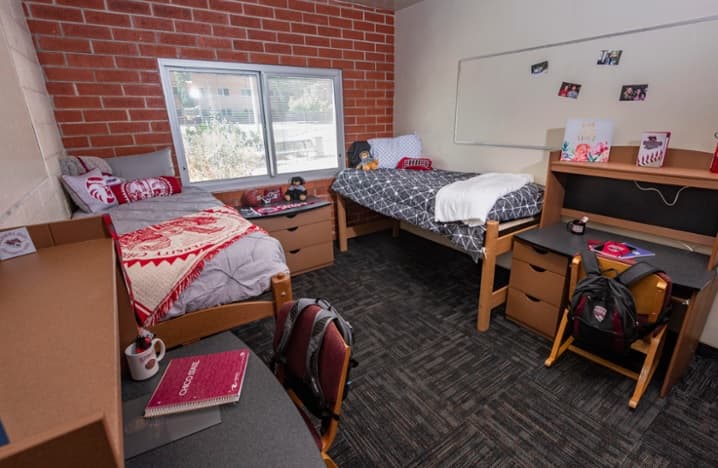 A dorm room with two beds, each with bedding and pillows, two desks with chairs and backpacks, a window, a whiteboard with photos, and personal items on the desks and dresser.