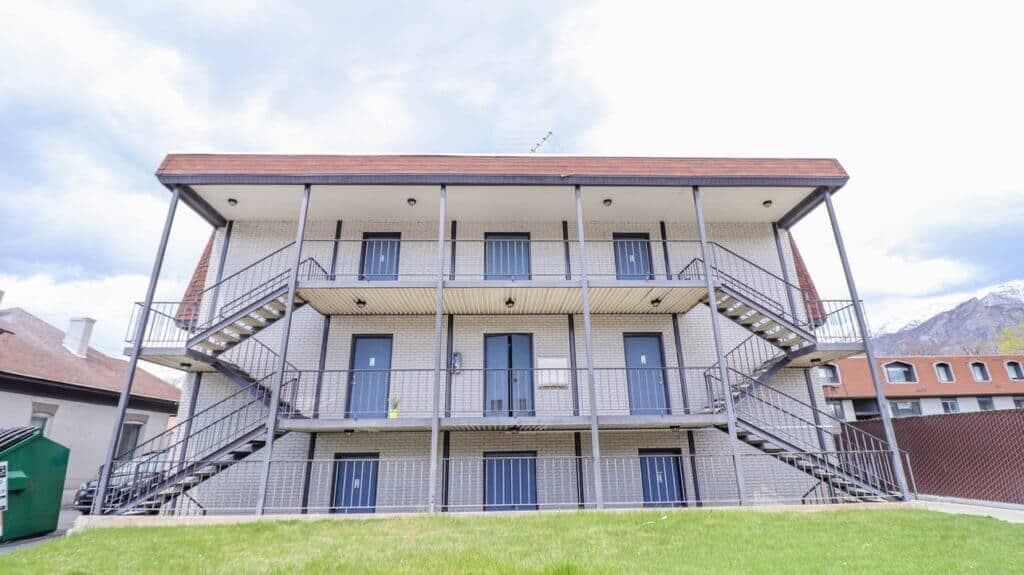 Liberty on Eighth Provo features a two-story apartment building with exterior stairs on each side, metal railings, and brick walls. Several doors line each level, and a grassy area stretches out front beneath the cloudy sky.