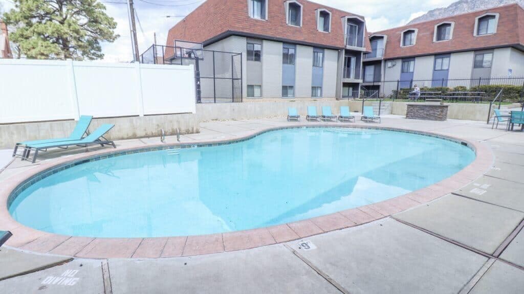 An outdoor kidney-shaped swimming pool at Liberty on Eighth Provo is surrounded by a concrete patio, lounge chairs, and residential apartment buildings in the background under a partly cloudy sky.
