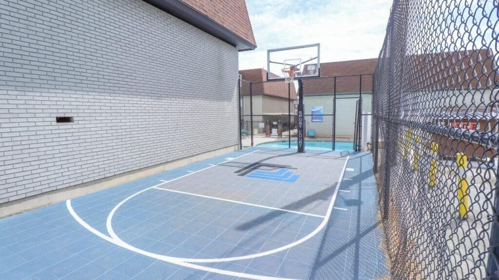 A small outdoor basketball court with a blue-gray surface, white lines, and a single hoop sits at Liberty on Eighth Provo, surrounded by a tall chain-link fence and bordered by light brick buildings.