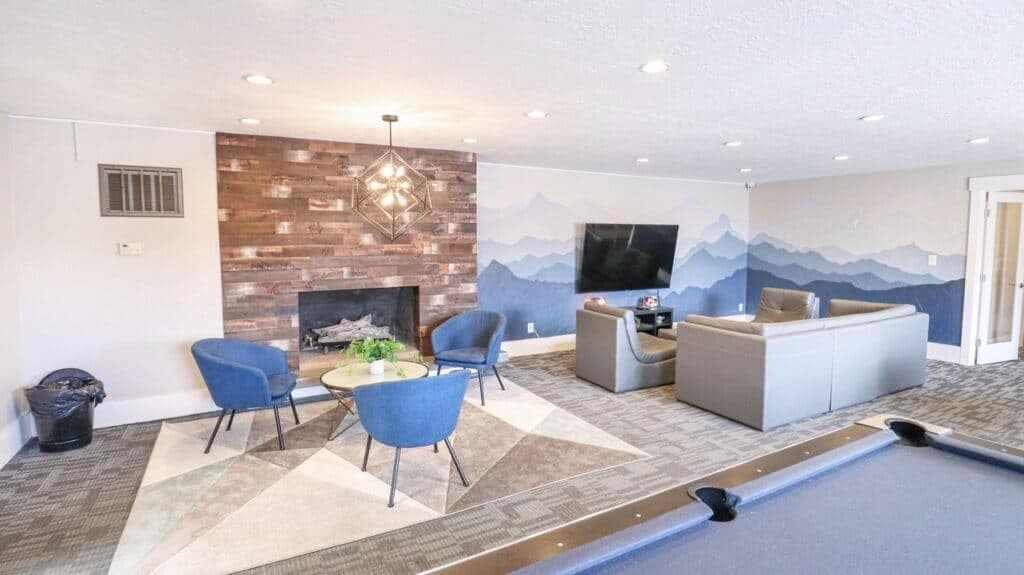 A modern lounge at Liberty on Eighth Provo features blue armchairs around a small table, a gray sofa set near a large TV, a wooden accent wall with fireplace, mountain mural, and part of a pool table in the foreground.