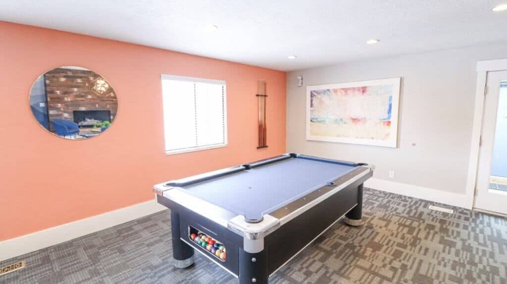A modern room at Liberty on Eighth Provo features a pool table with colorful balls, an orange accent wall and round mirror, large window, abstract artwork, and patterned gray carpet.