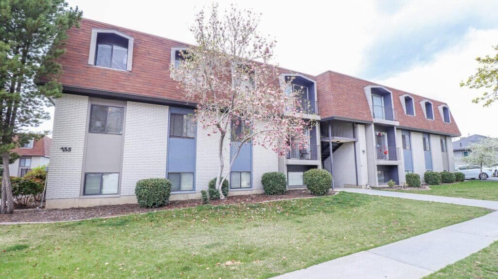 Liberty on Eighth Provo features a two-story apartment building with a brown roof, white and blue exterior walls, and multiple windows. Neatly trimmed bushes, a leafless tree, and a sidewalk accent the grassy lawn in front.