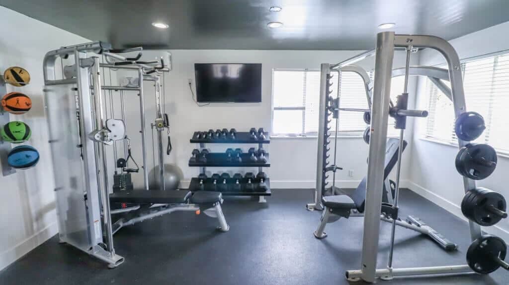 The small, modern gym at Liberty on Eighth Provo features weight machines, a bench, free weights on racks, medicine balls mounted on the wall, and a flat-screen TV. Large windows fill the space with natural light.