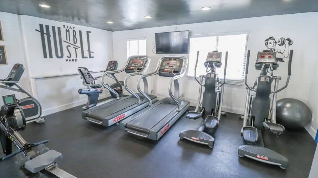 A small gym at Liberty on Eighth Provo features treadmills, elliptical machines, a rowing machine, a stability ball, inspiring wall art saying "Stay humble hustle hard," windows for natural light, and a TV mounted on the wall.