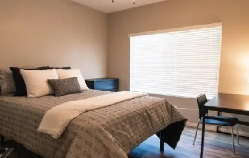 A neatly made bed with patterned bedding is next to a large window with blinds at Lofts on 8th Tempe. A dark desk with a chair sits by the window, and the room features beige walls and wooden floors.