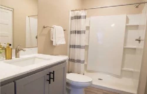 A modern bathroom at Lofts on 8th Tempe features a white sink, gray vanity, large mirror, towel rack with white towels, toilet, and a walk-in shower with a striped curtain and white walls.