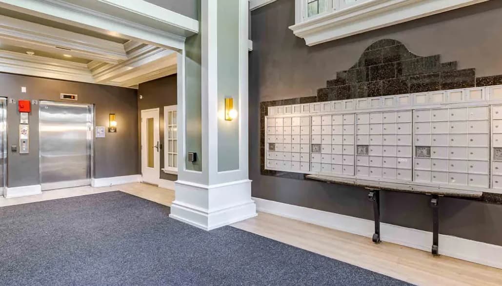 Apartment lobby in Yugo Seattle Lothlorien student housing, featuring gray walls, mailboxes along one wall, an elevator with metallic doors, white trim, overhead lighting, and blue carpeted flooring.