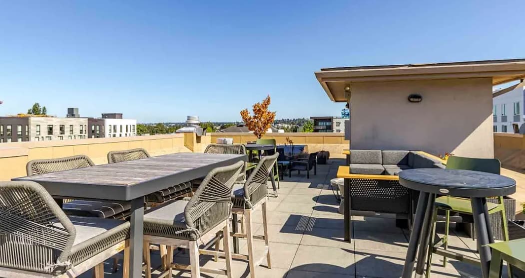 A rooftop patio at Yugo Seattle with modern tables, chairs, and sofas offers outdoor seating around a small tree, surrounded by city buildings under a clear blue sky—perfect for relaxing in Seattle student housing.
