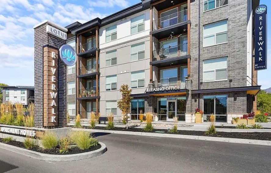 Modern apartment building exterior with a Riverwalk sign and The Lotus Boulder nearby, featuring a Leasing Office entrance, multiple balconies, large windows, and landscaped areas with ornamental grasses and shrubs along the driveway.