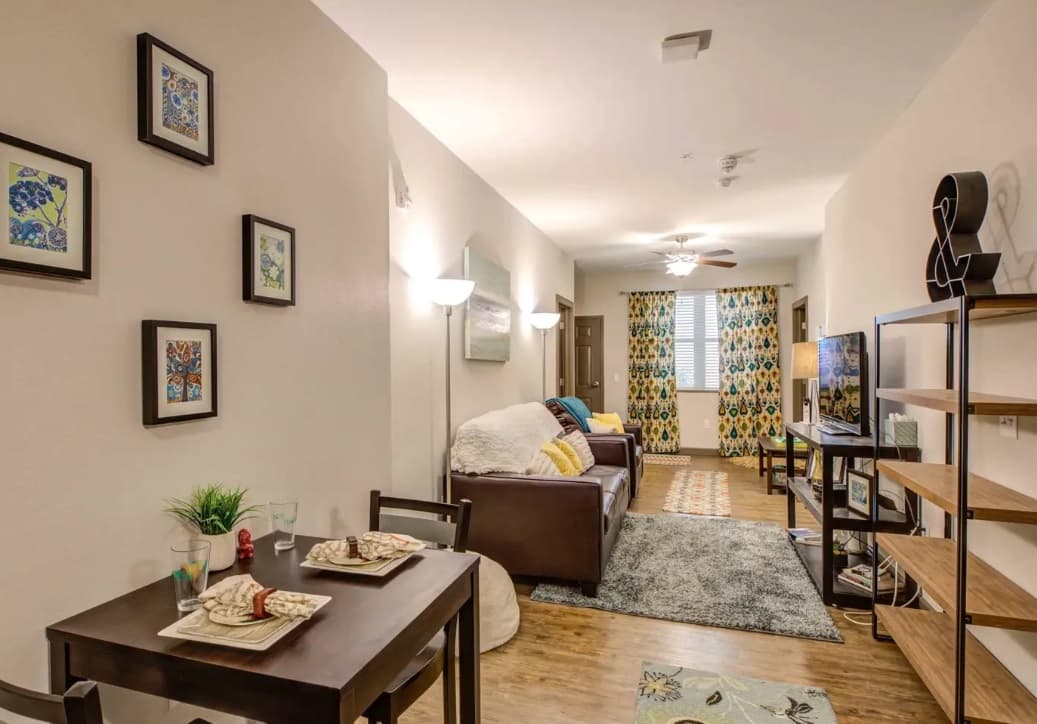 A cozy living and dining area in The Lotus Boulder apartment features a small table set for two, a brown sofa with colorful pillows, a TV, shelves, wall art, and patterned curtains. Warm lighting creates a welcoming atmosphere.