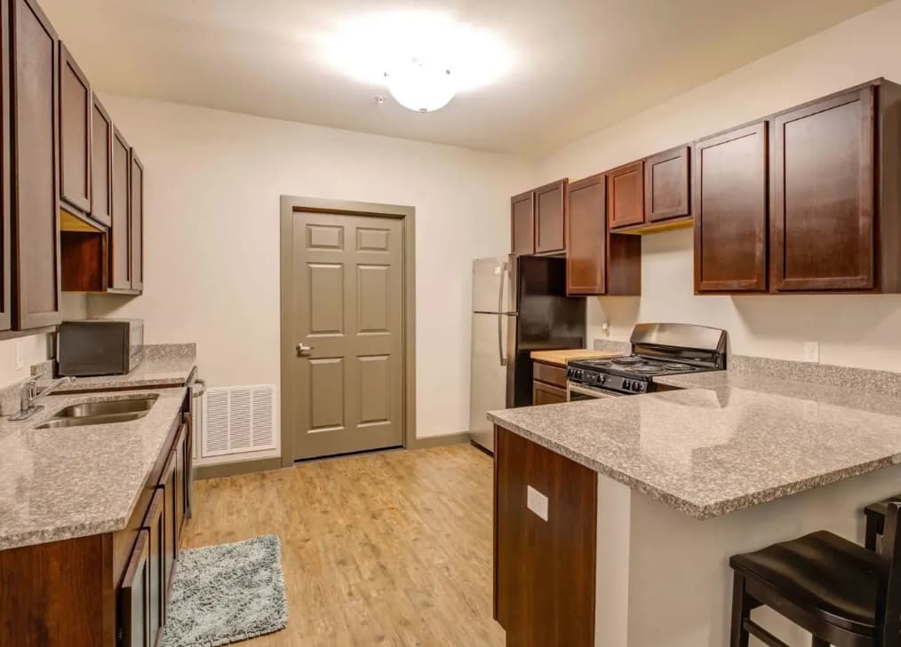 Modern kitchen at The Lotus Boulder with wood cabinets, granite countertops, stainless steel appliances, wooden floor, central light fixture, a door at the far end, and two bar stools at the counter on the right.