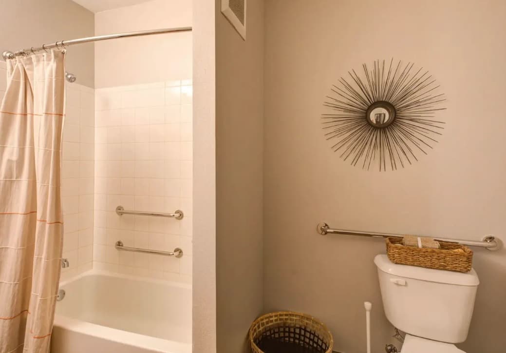 A bathroom at The Lotus Boulder features a white bathtub and shower, grab bars, a toilet with more grab bars, a woven basket on top, a laundry basket on the floor, and a decorative sunburst wall mirror above the toilet.
