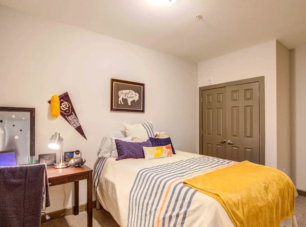 A cozy bedroom with a striped bedspread, yellow throw, and multiple pillows. A desk with a lamp and books sits to the left, while The Lotus Boulder pennant and bison art add charm above the bed. There’s a double-door closet.