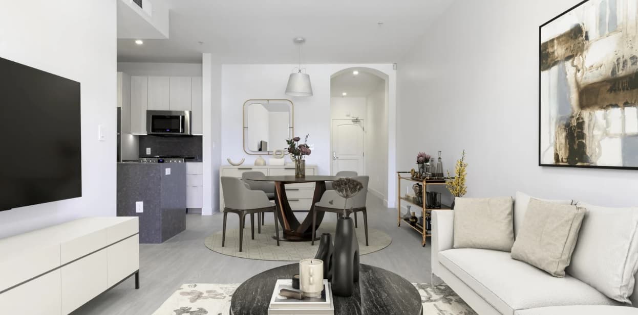 Modern living and dining area with a white sofa, round coffee table, wall art, flat-screen TV, and a dining table for four near a kitchen with white cabinets and a microwave. Neutral tones and minimalist decor throughout.