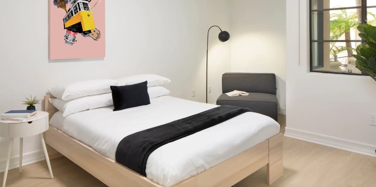 Minimalist bedroom with a wooden bed, white bedding, black accent pillow and blanket, small round nightstand, gray chair, modern floor lamp, large window, plant, and colorful wall art.