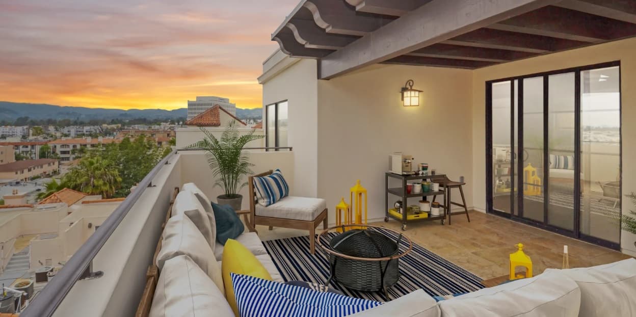 A modern rooftop patio at sunset features a white sectional sofa, striped cushions, a glass door, and city views. A rug, chairs, and yellow lanterns decorate the cozy outdoor space.