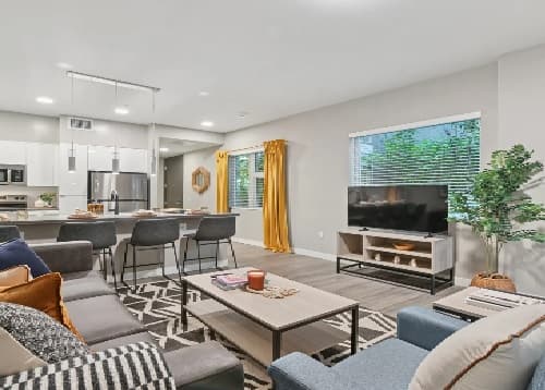Modern living room at The Mark Los Angeles with a sectional sofa, patterned rug, coffee table, and TV on a stand. A large window with yellow curtains brightens the space, while the kitchen with bar stools and a potted plant add style.