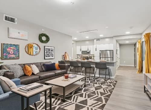 A modern, open-concept living room and kitchen by Mark Los Angeles features a sectional sofa, accent pillows, patterned rug, wall art, bar stools at a kitchen island, stainless steel appliances, and bright neutral decor with pops of yellow.