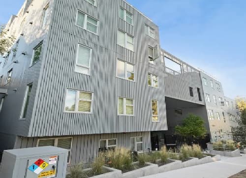 A modern, multi-story apartment building—The Mark Los Angeles—features striped gray metal siding and many windows. A small landscaped area with grass and shrubs sits in front, along with a utility box on the sidewalk beneath a clear blue sky.