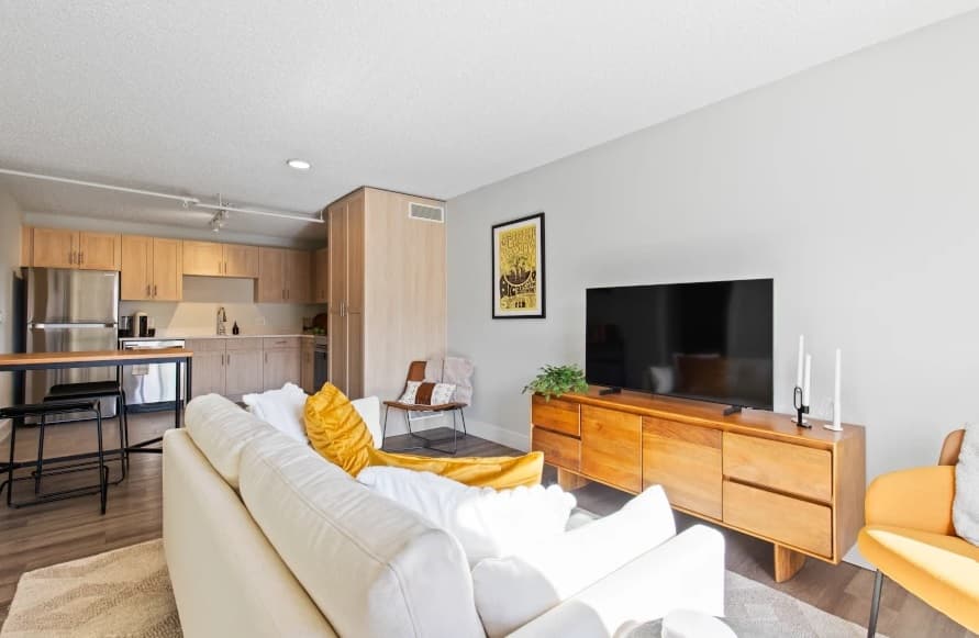 A modern, sunlit living room in Mountainaire Boulder features a white sofa with yellow pillows, a wooden TV stand with a large flat-screen TV, and an open kitchen with light wood cabinets and stainless steel appliances in the background.